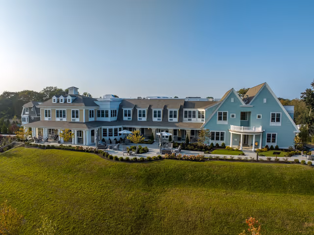 A large, multi-story senior living facility building with a light blue and beige exterior, multiple windows, and a spacious patio area with seating. The building is surrounded by a well-maintained lawn and landscaping under a clear blue sky.