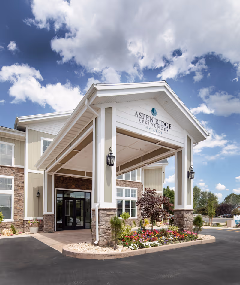 Front entrance of Aspen Ridge Residences of Lehi, showing a covered driveway with stone and beige siding, decorative lanterns, and a landscaped flower bed under a partly cloudy sky.