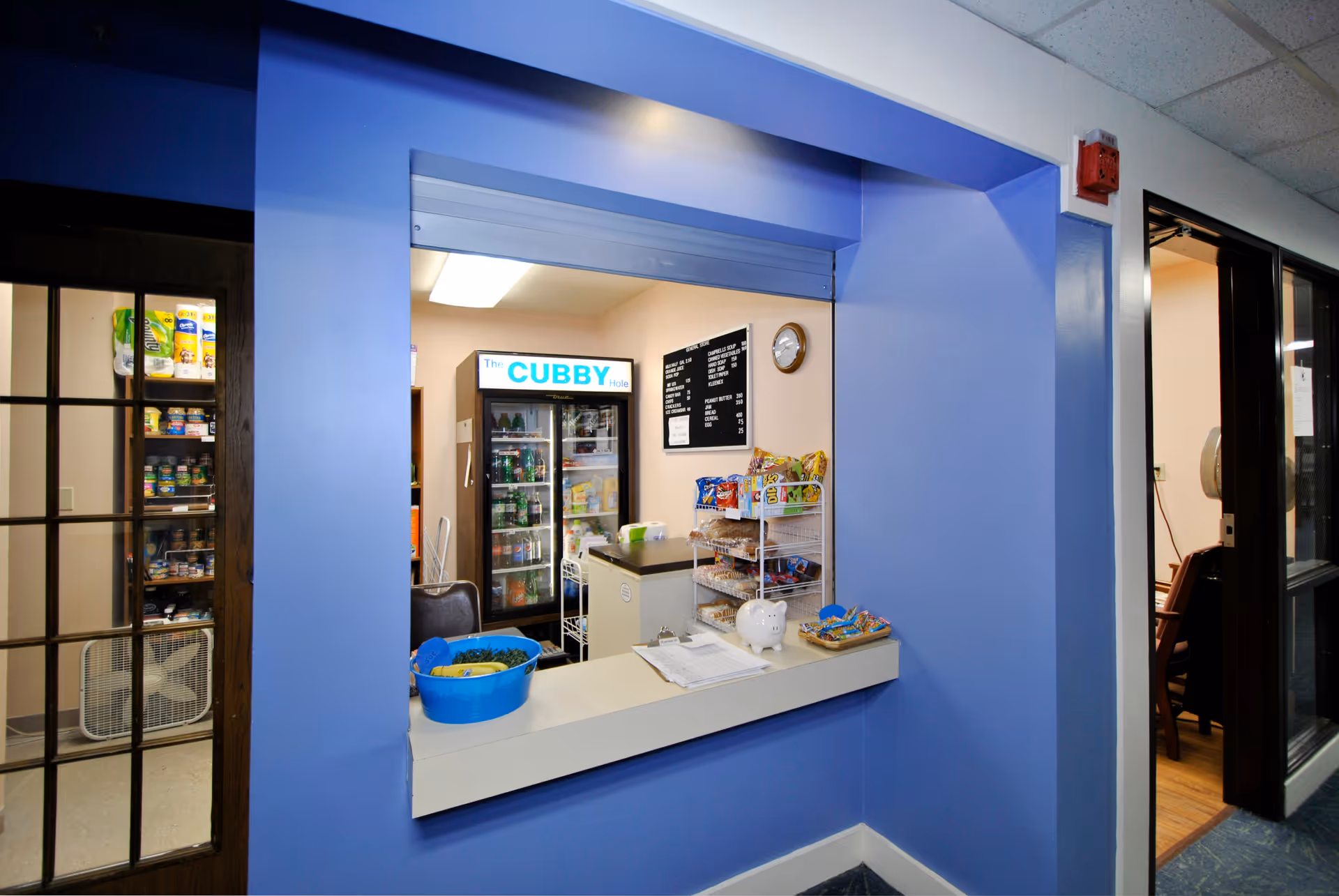 A small snack and beverage area named 'The Cubby Hole' with a counter window framed by blue walls. Inside, there is a refrigerator stocked with drinks, shelves with snacks, a clock on the wall, and a menu board. To the left, a glass door reveals shelves stocked with various canned goods and a fan on the floor. To the right, an open doorway leads to a room with chairs and a wooden floor.