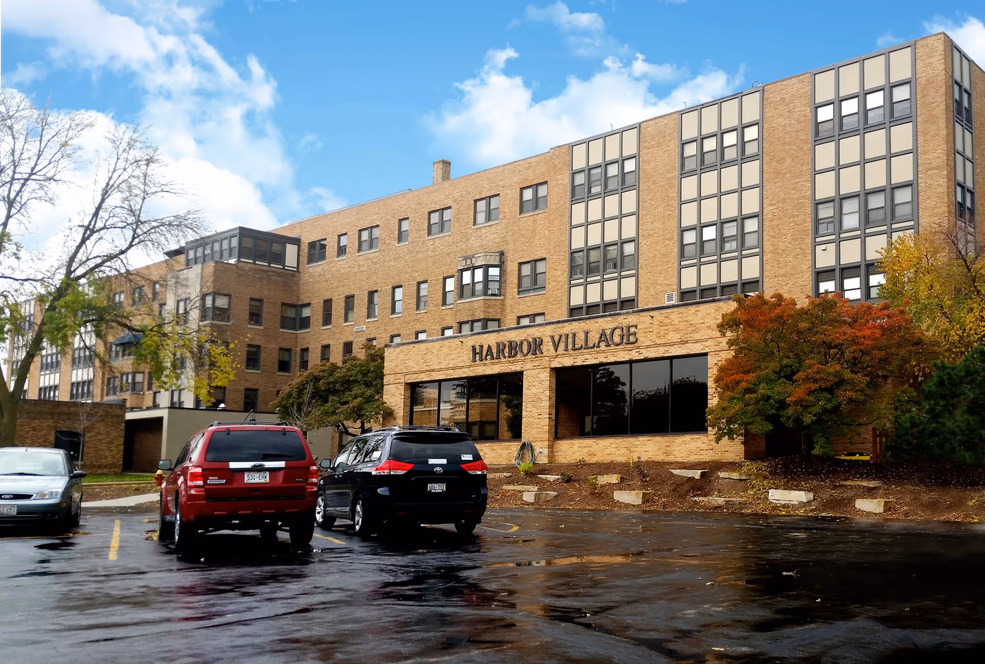 Exterior view of a multi-story brick building with the sign 'Harbor Village' on the front. There are several windows and a few trees with autumn foliage around the building. In the foreground, there is a parking lot with three parked cars under a partly cloudy sky.