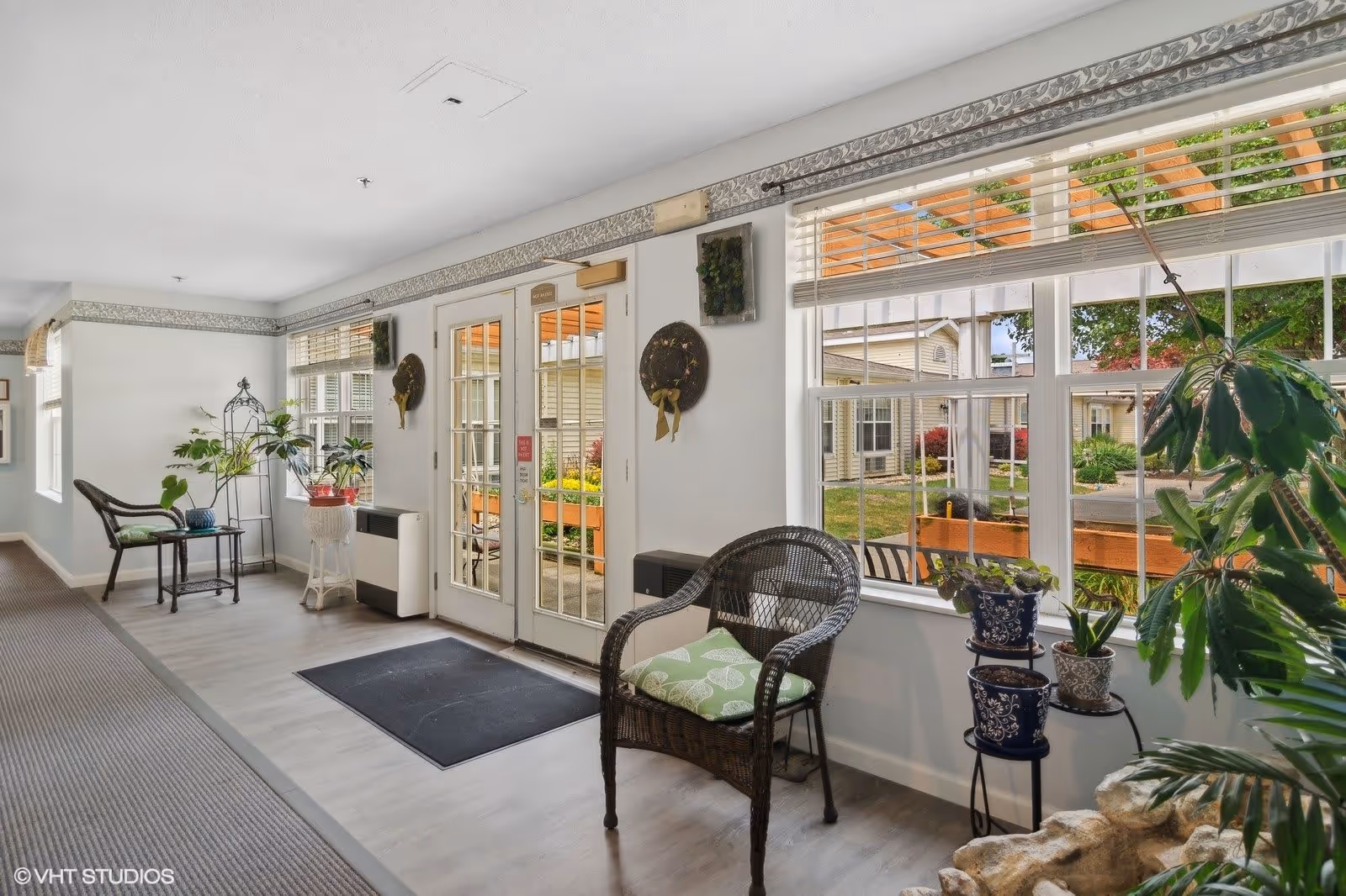 Bright interior sitting area with wicker chairs, potted plants, and French doors and large windows overlooking a garden.