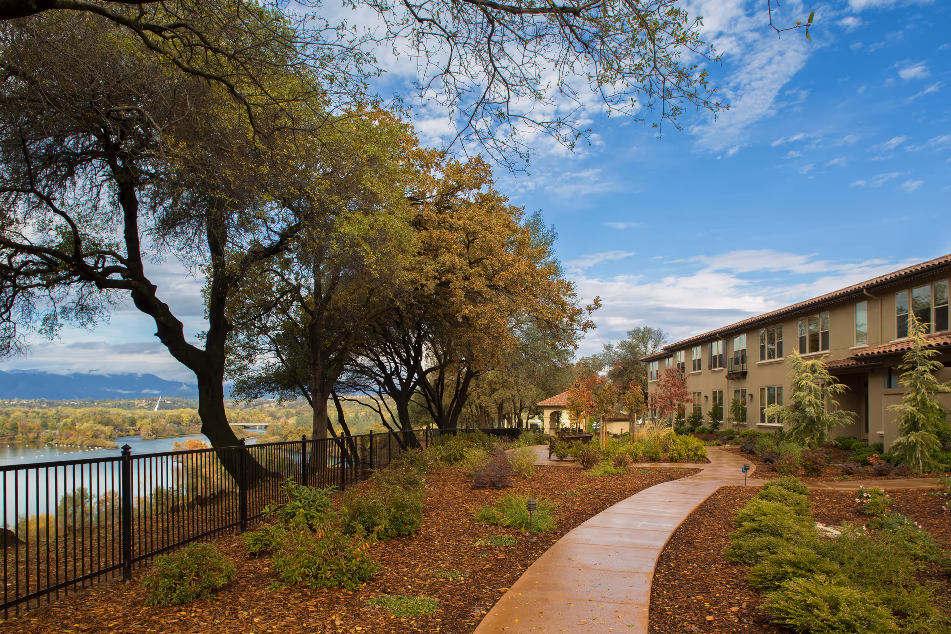 A winding paved pathway through a landscaped garden area with various shrubs and trees, leading to a two-story building with multiple windows. A black metal fence runs along the left side, overlooking a body of water and distant mountains under a partly cloudy blue sky.