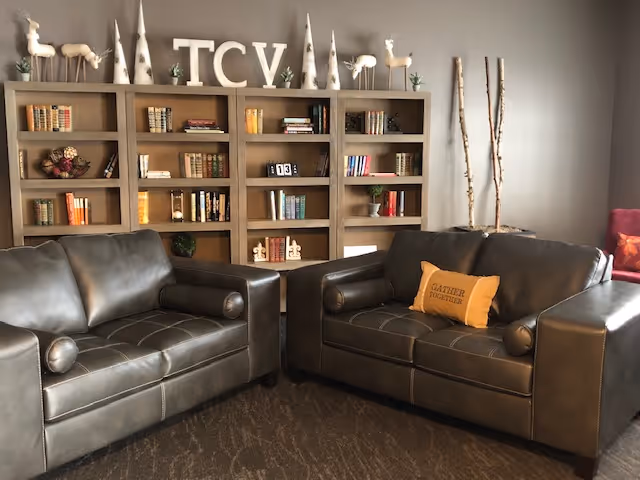 A cozy living room area with two dark leather sofas facing each other. Behind the sofas is a wooden bookshelf filled with books and decorative items, including letters spelling 'TCV' on top. The room has a neutral color palette with a carpeted floor and some tall decorative sticks in the corner.