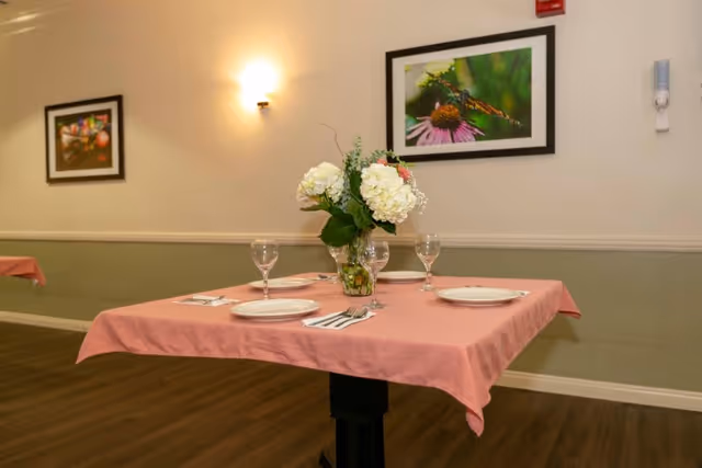 A small dining table with a pink tablecloth set with plates, wine glasses and a floral centerpiece in a dining room with framed nature photos on the wall.