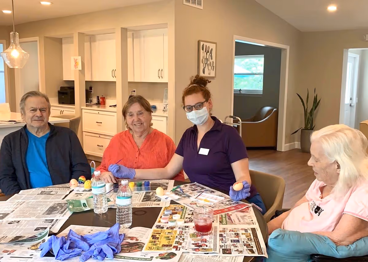 A masked caregiver and three senior residents sit around a table covered with newspapers and craft supplies in a communal room.