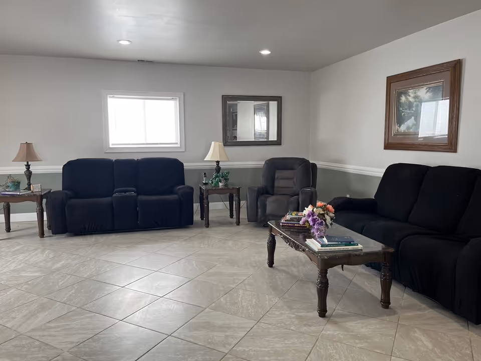 A living room with two black sofas and a black recliner chair arranged around a wooden coffee table with books and a flower arrangement. There are two side tables with lamps, a window, a mirror, and a framed picture on the walls. The floor is tiled with light-colored tiles.