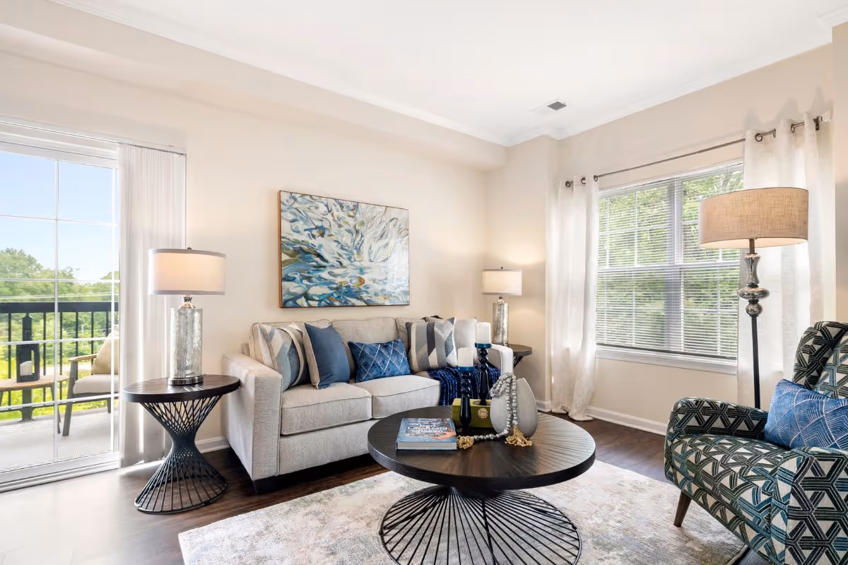 Bright living room with a sofa, patterned armchair, round coffee table, lamps, artwork, and a sliding door leading to a balcony.