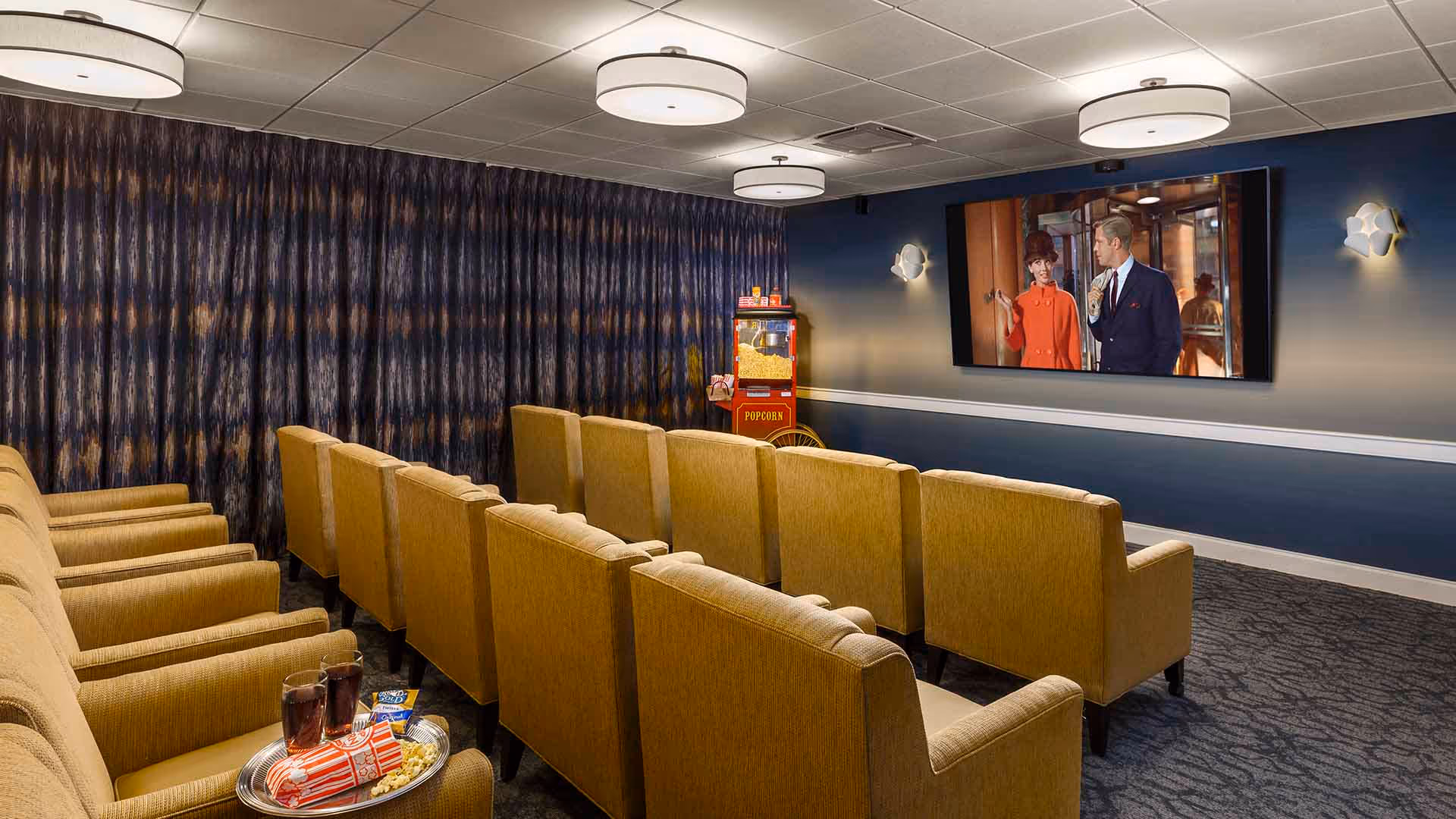 A small movie theater room with two rows of mustard yellow upholstered armchairs facing a large flat-screen TV mounted on a blue wall. A popcorn machine is placed in the corner near the TV. The room has dark patterned curtains along one wall and round ceiling lights providing illumination.