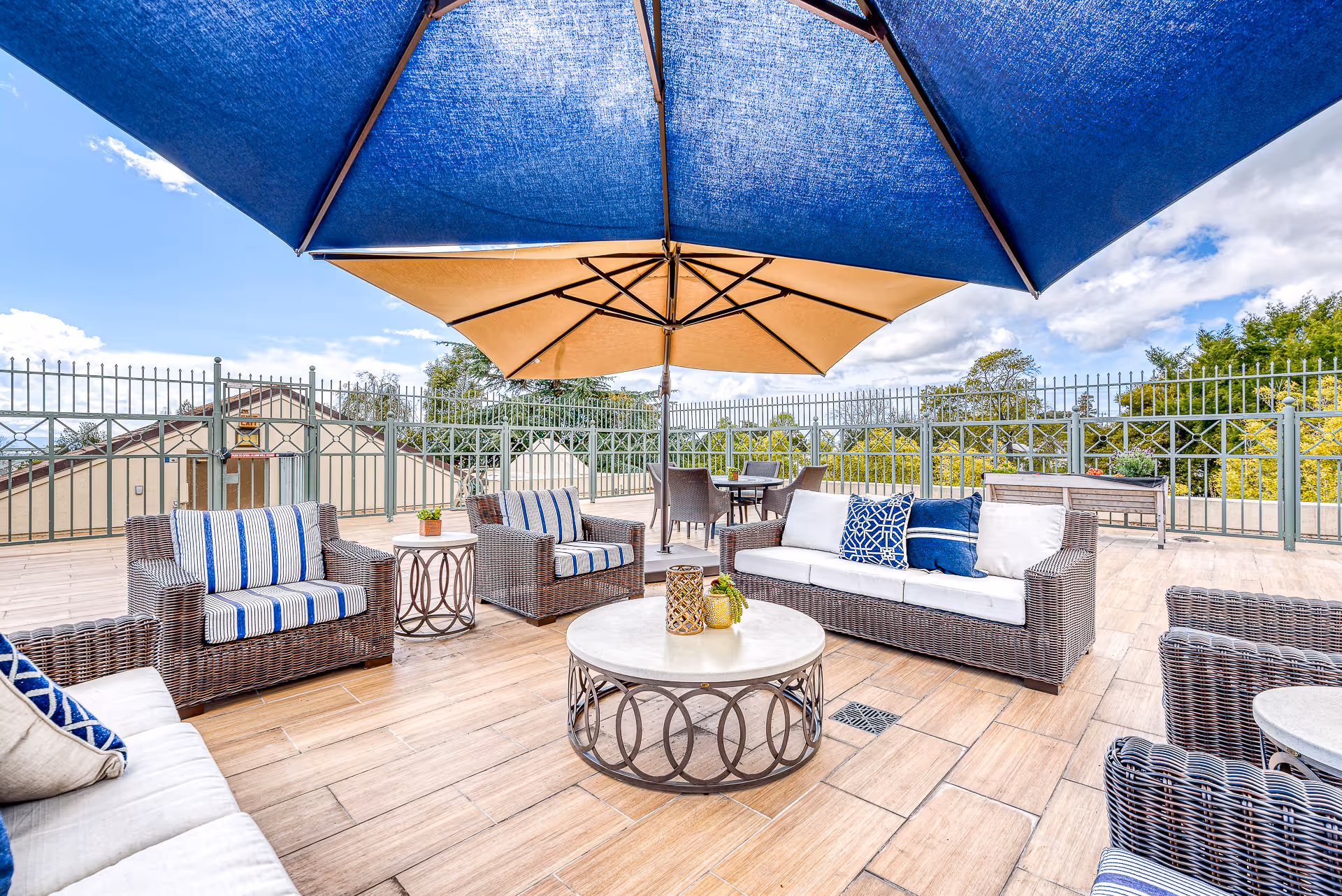 Outdoor patio area with wicker furniture including sofas and armchairs with cushions, a round coffee table, and large blue and beige umbrellas providing shade. The patio is surrounded by a metal fence and there are trees and a partly cloudy sky in the background.