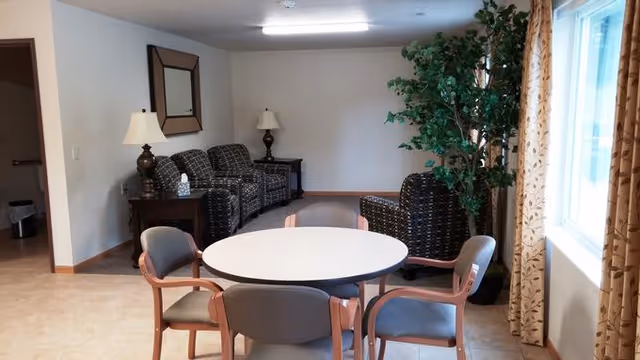 A cozy common area with a round white table surrounded by four chairs with wooden frames and gray cushions. In the background, there are three patterned armchairs, two side tables with lamps, a large mirror on the wall, and a tall green potted plant near a window with beige curtains featuring a leaf pattern.