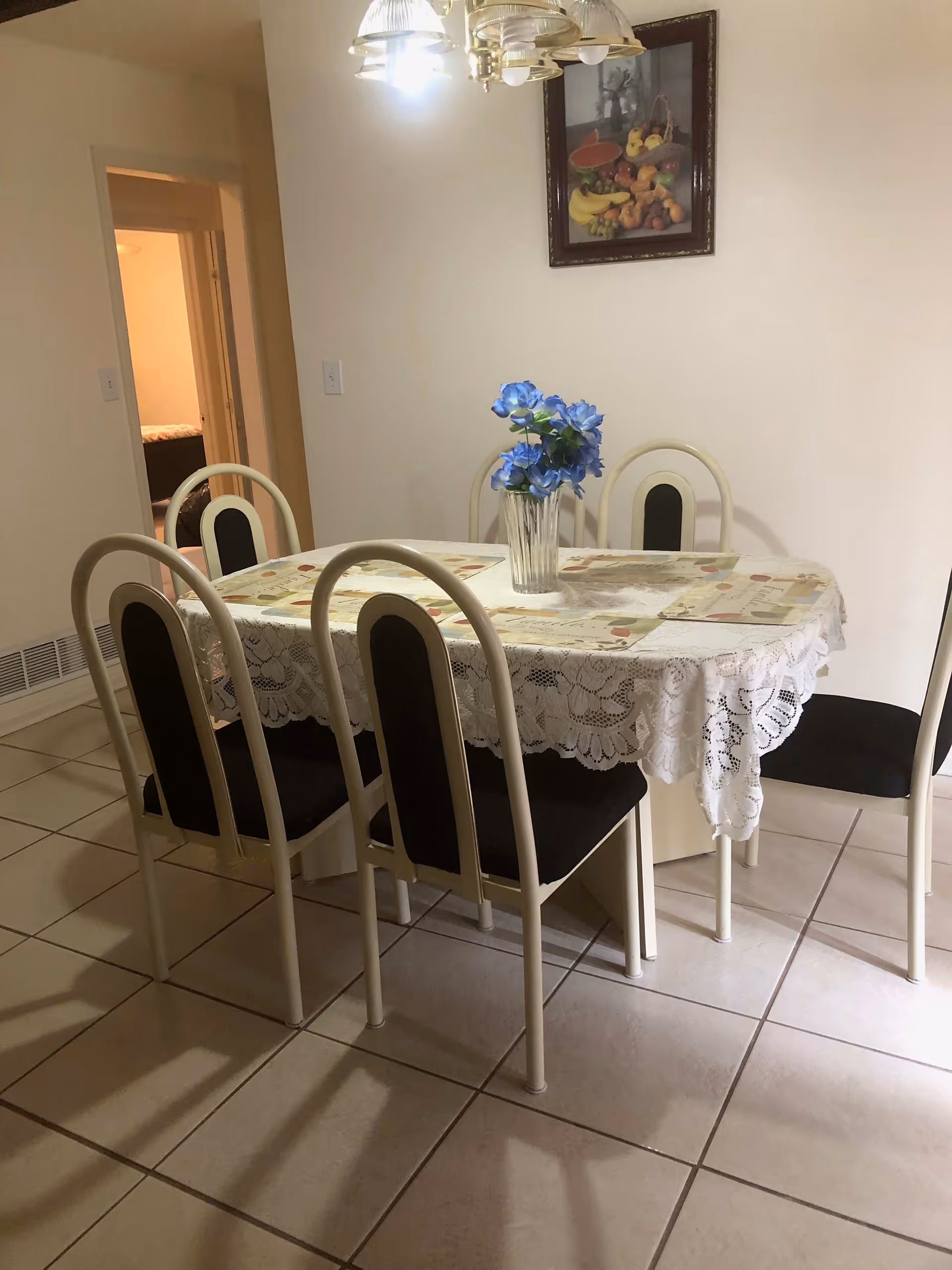 A dining room with a rectangular table covered with a lace tablecloth and placemats. There are six chairs with black cushions around the table. A vase with blue flowers is placed in the center of the table. A framed picture of fruit hangs on the wall above the table. The room has tiled flooring and a doorway leading to another room is visible in the background.