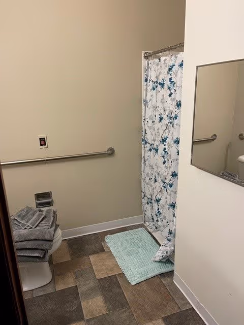 A bathroom with a tiled floor, a shower area with a floral patterned curtain, a light blue bath mat, a wall-mounted grab bar, a mirror on the right wall, and a stack of folded gray towels on a white surface.