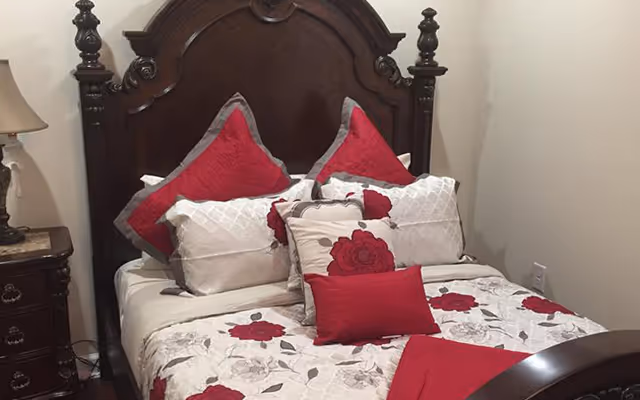 A bedroom with a dark wooden bed frame featuring an ornate headboard. The bed is made with white bedding decorated with red and gray floral patterns, and several pillows in matching colors are arranged neatly. A wooden nightstand with a lamp is visible on the left side of the bed.