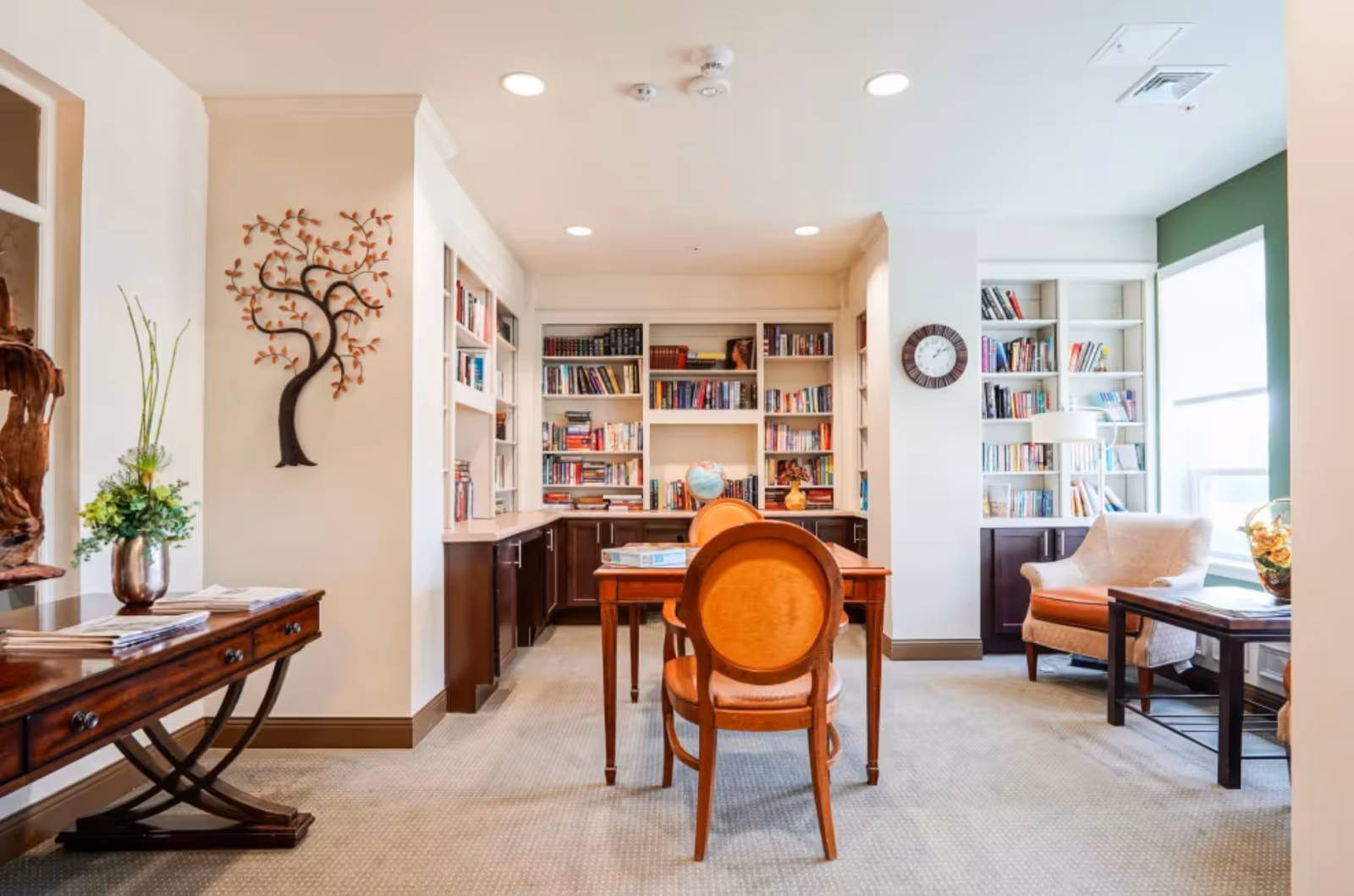 Well-lit communal library room with built-in bookshelves, a central table and chairs, and armchairs by a window.
