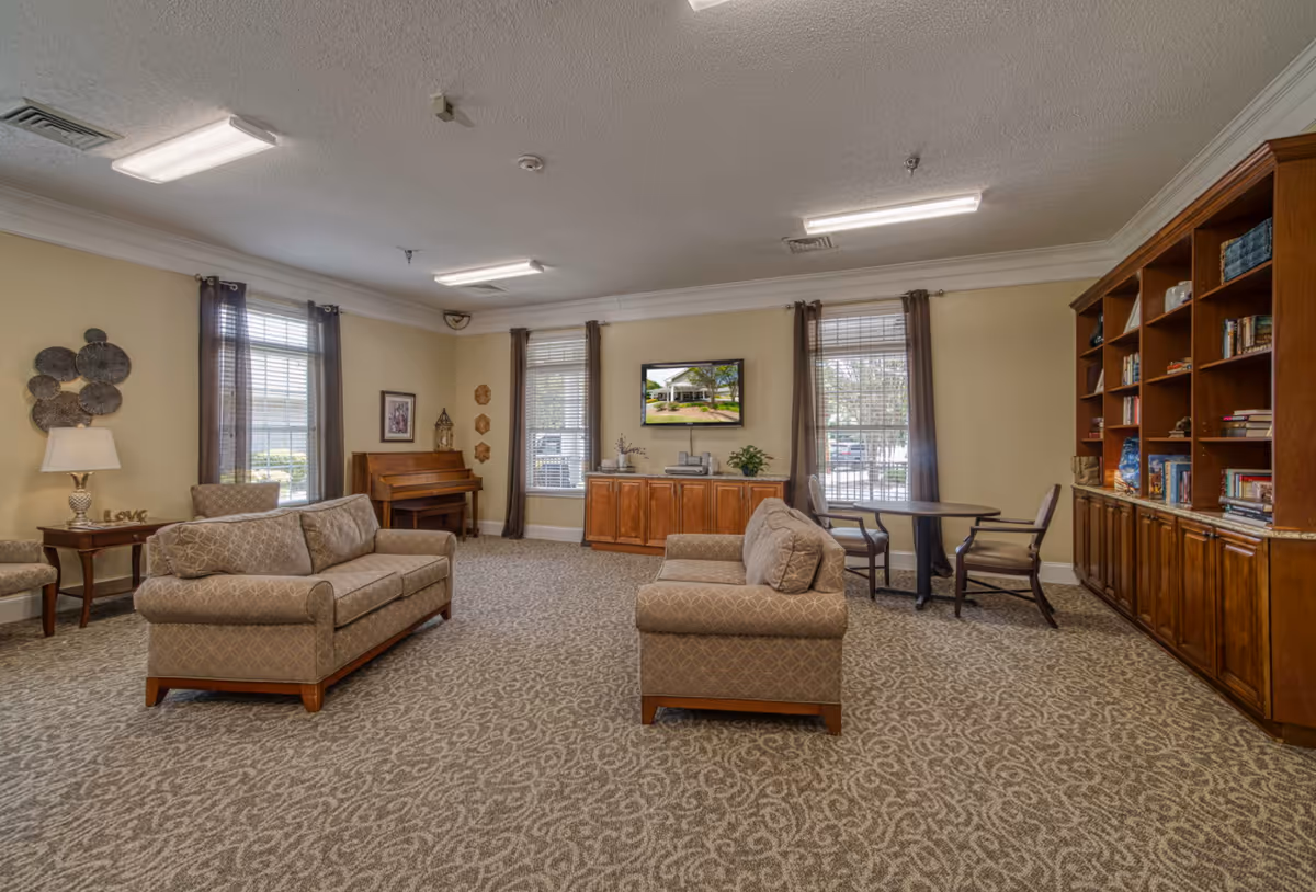A spacious living room with two beige patterned sofas facing each other on a patterned carpet. The room has three large windows with brown curtains, a wooden piano in the corner, a wooden cabinet with a TV mounted above it, and a round table with two chairs. A large wooden bookshelf filled with books and decorative items lines one wall.