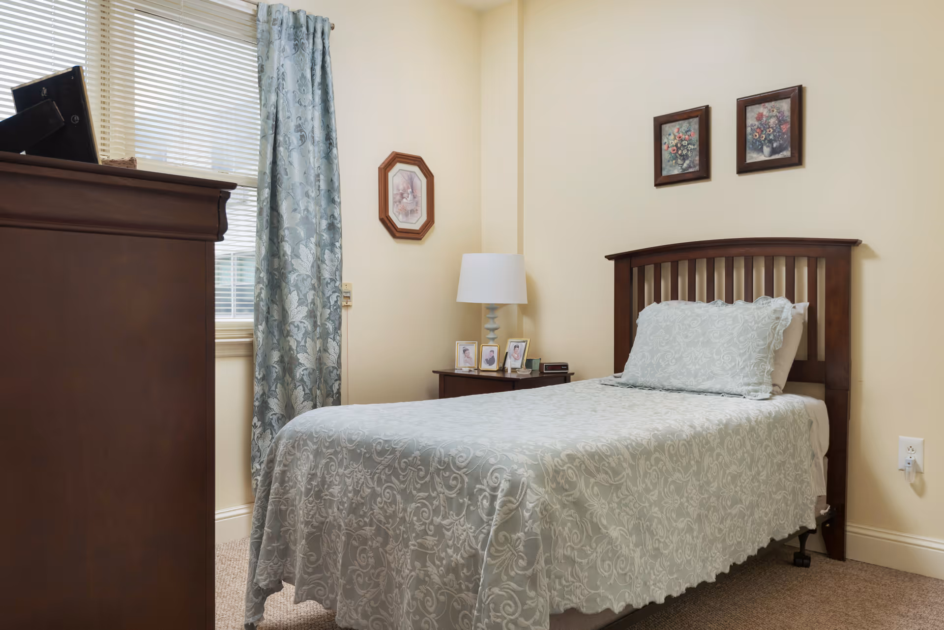 A neatly made single bed with a light blue patterned bedspread and matching pillow in a bedroom. Next to the bed is a wooden nightstand with a white lamp and framed photos. The room has beige walls with three framed floral pictures hanging above the bed. A window with blinds and blue patterned curtains is visible behind a dark wooden dresser.