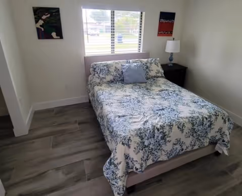 Bedroom with a floral bedspread on a double bed, a window with blinds, a nightstand with a lamp, and wall art.