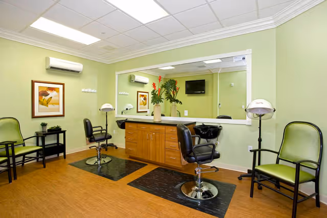 Interior view of a salon area in a senior living facility with two black salon chairs in front of a large mirror, a wooden cabinet with drawers, two hair dryers on stands, green walls, two green chairs along the wall, a small black side table with a plant, and a framed picture on the wall.