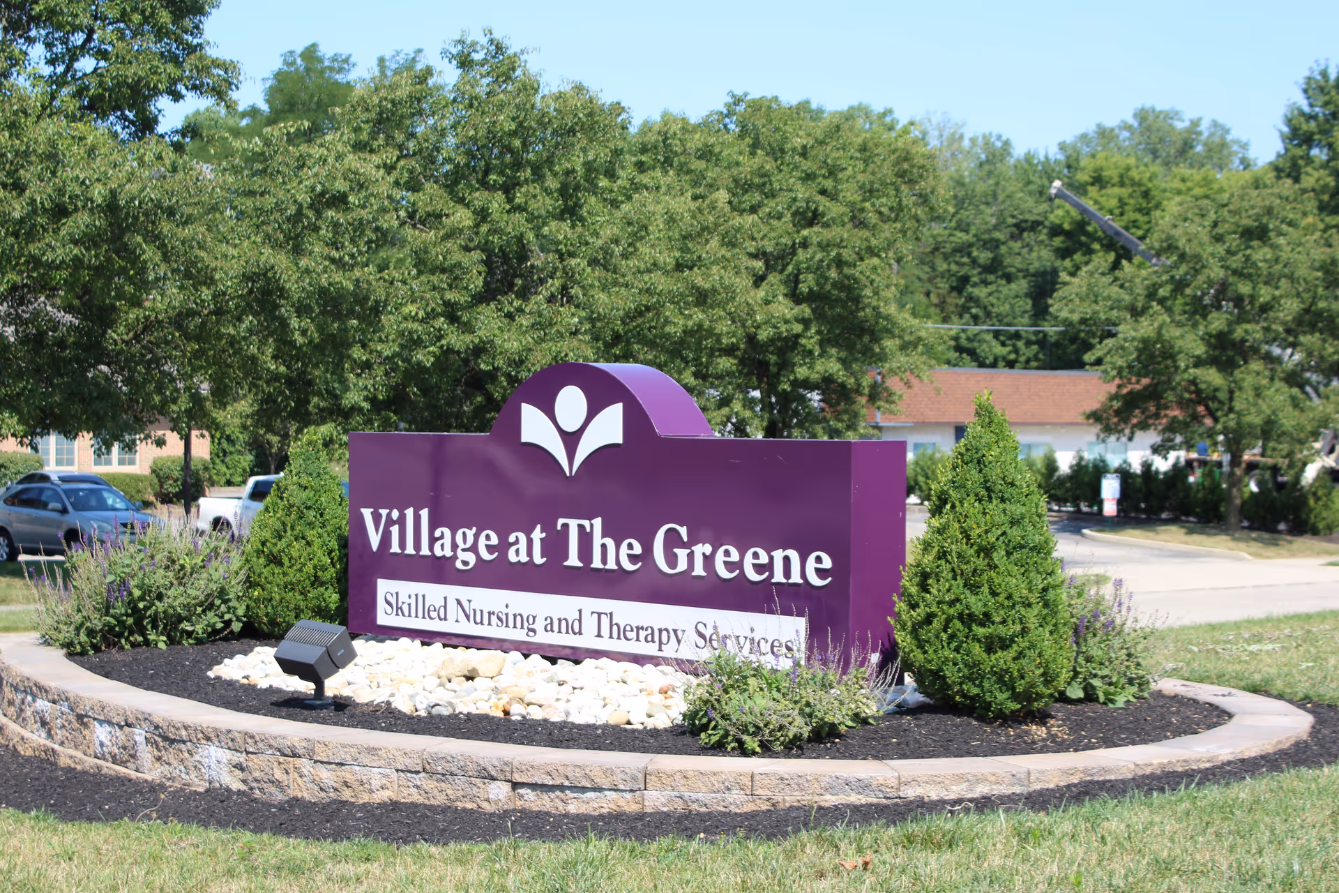 Outdoor view of a purple sign for Village at The Greene, a skilled nursing and therapy services facility, surrounded by landscaping with bushes, flowers, and rocks, with trees and parked cars in the background.