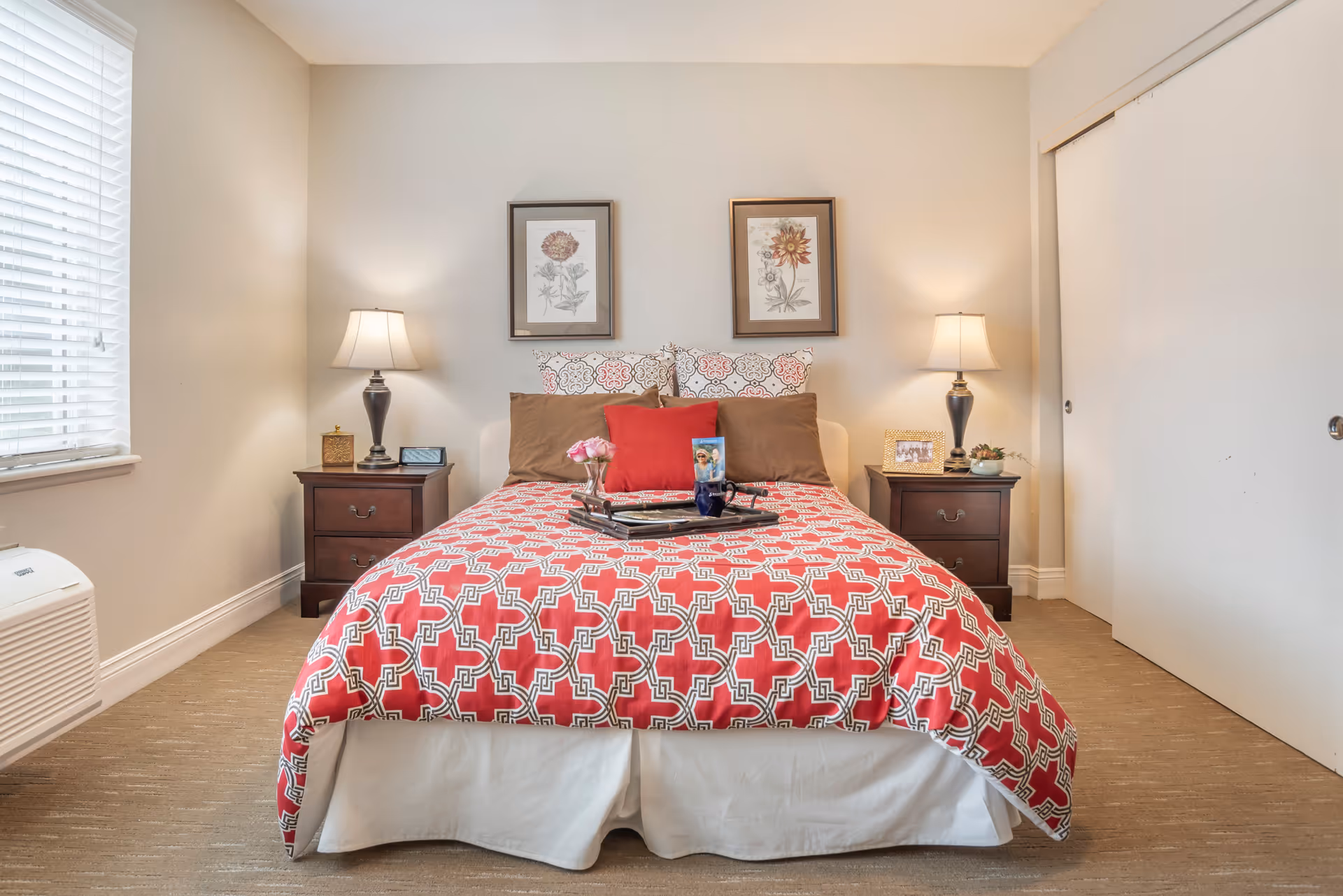 A neatly made bed with a red and white patterned comforter and multiple pillows, flanked by two dark wooden nightstands each with a lamp. Above the bed are two framed floral prints. There is a window with blinds on the left and a white sliding door closet on the right.