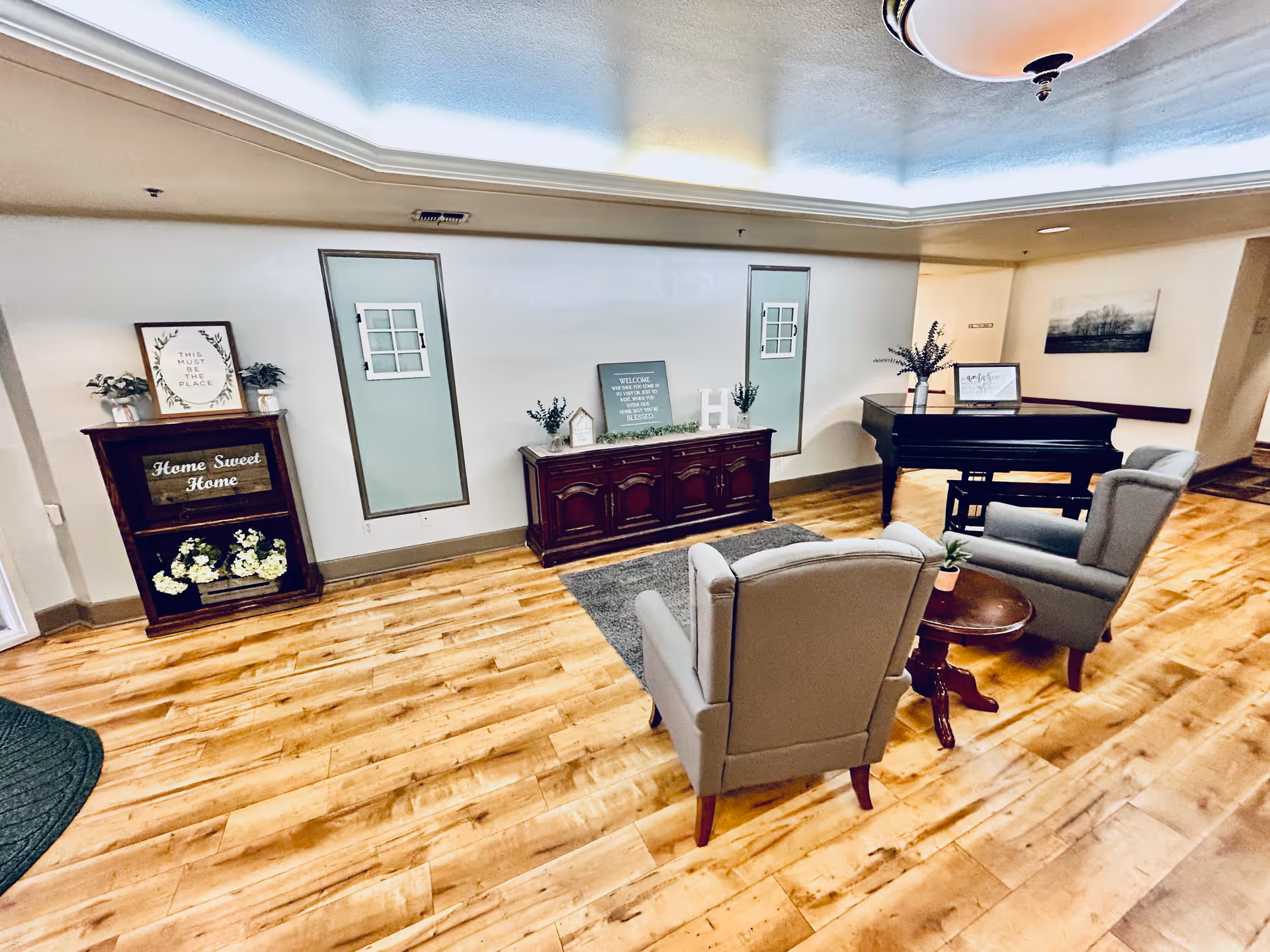 A cozy sitting area in a senior living facility with two gray armchairs around a small wooden table, a grand piano in the background, wooden flooring, and decorative furniture including a cabinet with plants and signs that say 'Home Sweet Home' and 'This Must Be The Place'.