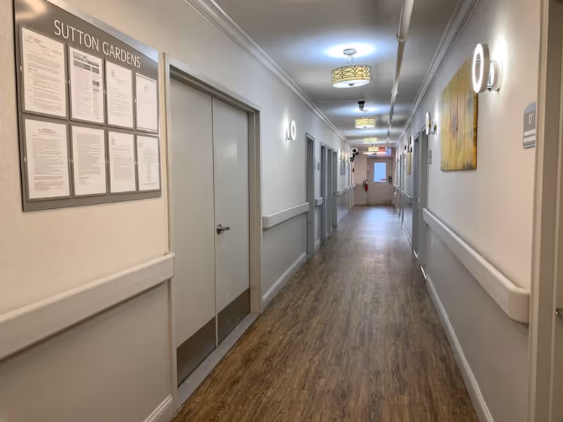 A long, well-lit hallway in a senior living facility named Sutton Gardens. The hallway has light-colored walls with handrails on both sides, wood-patterned flooring, and several closed doors along the corridor. There are framed notices on the left wall near the entrance and a piece of artwork on the right wall. Ceiling lights and wall sconces illuminate the space.