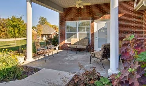 Covered outdoor patio area with brick walls, white columns, and outdoor seating including chairs and a loveseat. There are plants and greenery around the patio and a view of neighboring houses and trees in the background.