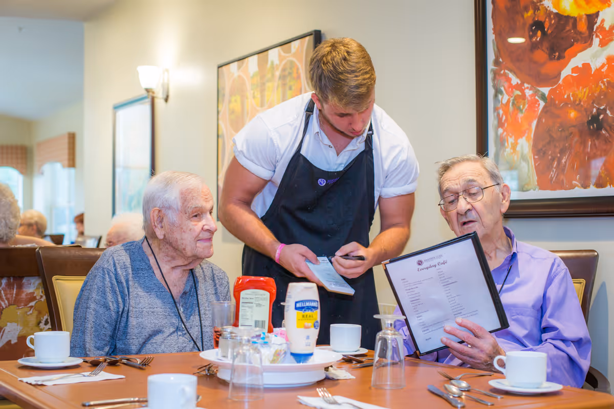 A waiter in a black apron takes an order from two elderly men seated at a dining table in a senior living facility. One man is looking at the menu while the other looks at the waiter. The table is set with cups, utensils, and condiments, and there is colorful artwork on the wall behind them.