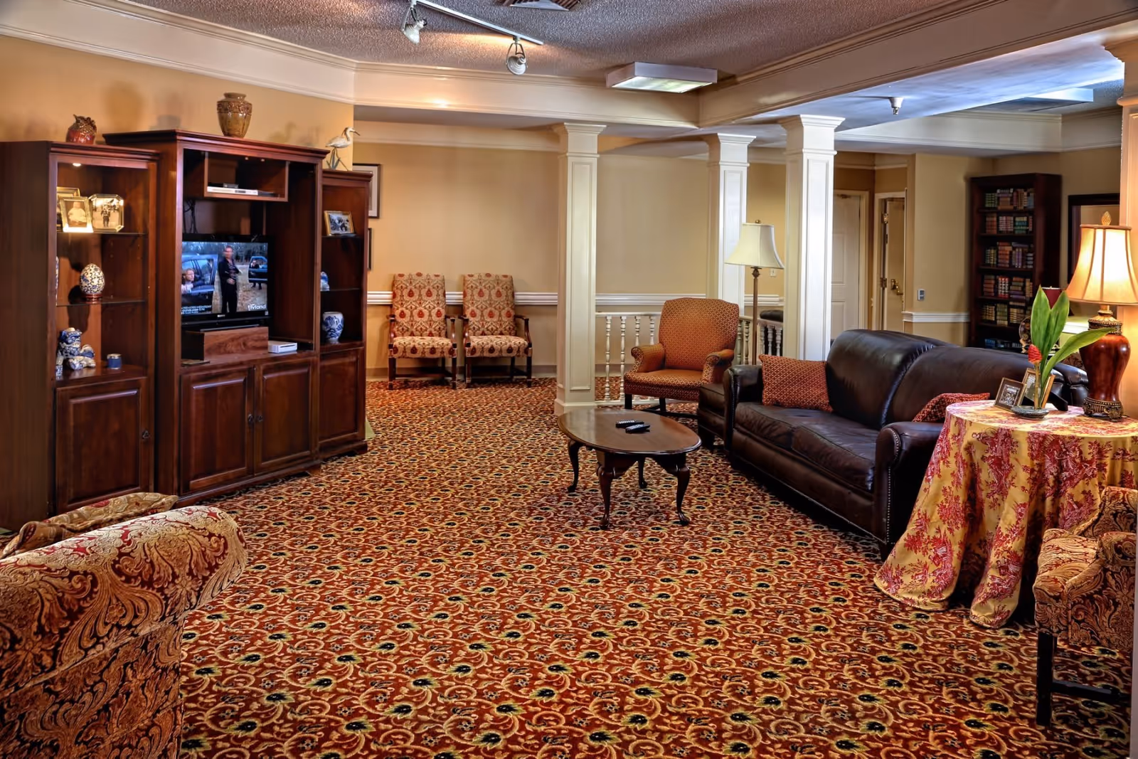 A cozy senior living facility common area with patterned red carpet, a dark brown leather sofa, an armchair, a wooden coffee table with remote controls, a wooden entertainment center with a TV, decorative items, and framed photos. There are two patterned chairs against the wall, a floor lamp, a table with a floral tablecloth and a potted plant, and a bookshelf filled with books in the background.