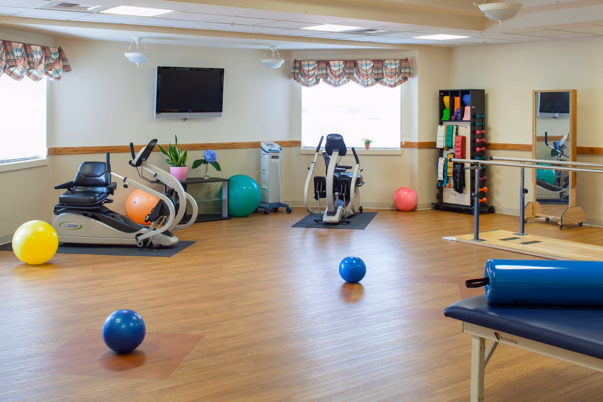 A bright and spacious exercise room with wooden flooring, featuring two stationary recumbent bikes, various colorful exercise balls scattered on the floor, a rack with small dumbbells and resistance bands, a wall-mounted TV, and a padded exercise table with a blue cylindrical cushion. The room has large windows with plaid valances allowing natural light to enter.