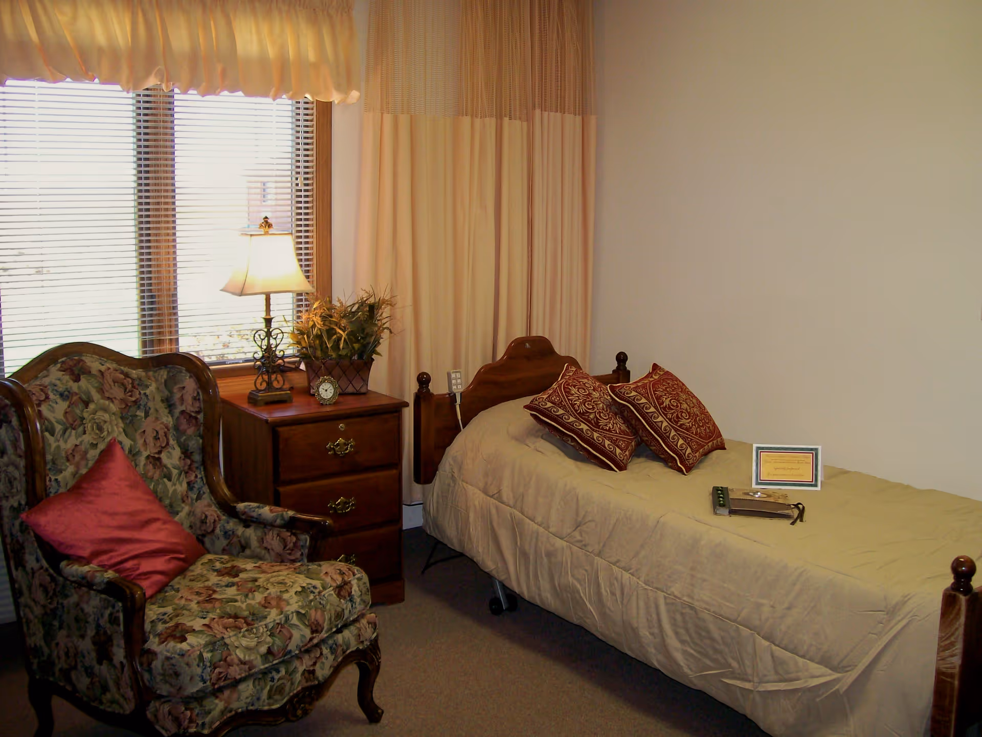 A cozy bedroom featuring a single bed with decorative pillows, a floral armchair, and a wooden nightstand with a lamp by a window.
