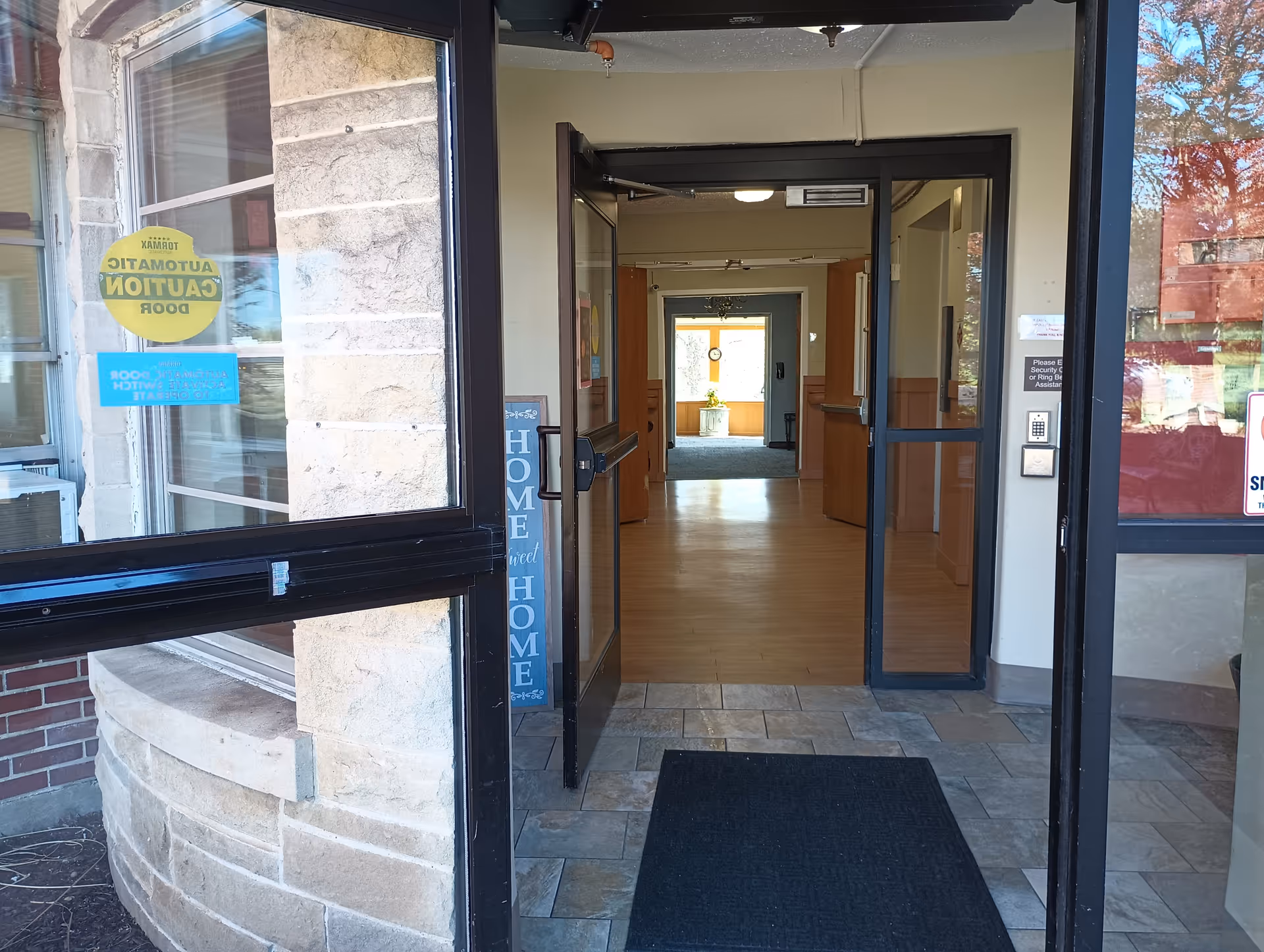 Entrance to a facility with glass doors open, showing a tiled floor mat at the entrance and a hallway leading to a room with a window and a clock on the wall. A sign near the door reads 'HOME sweet HOME'.