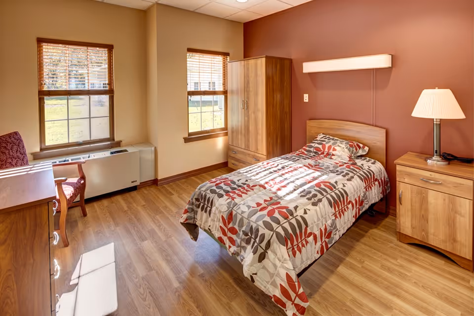 A bright and cozy bedroom in an assisted living residence featuring a single bed with a patterned bedspread in shades of red, gray, and white. The room has wooden flooring, two windows with wooden blinds letting in natural light, a wooden wardrobe, a nightstand with a lamp, a wooden desk, and a chair with a red patterned cushion.