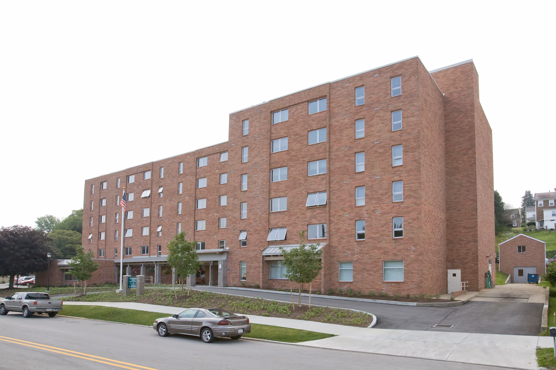 Exterior view of a multi-story brick building with several windows, a driveway, and a small landscaped area with trees and shrubs. Two cars are parked on the street in front of the building, and an American flag is visible near the entrance.