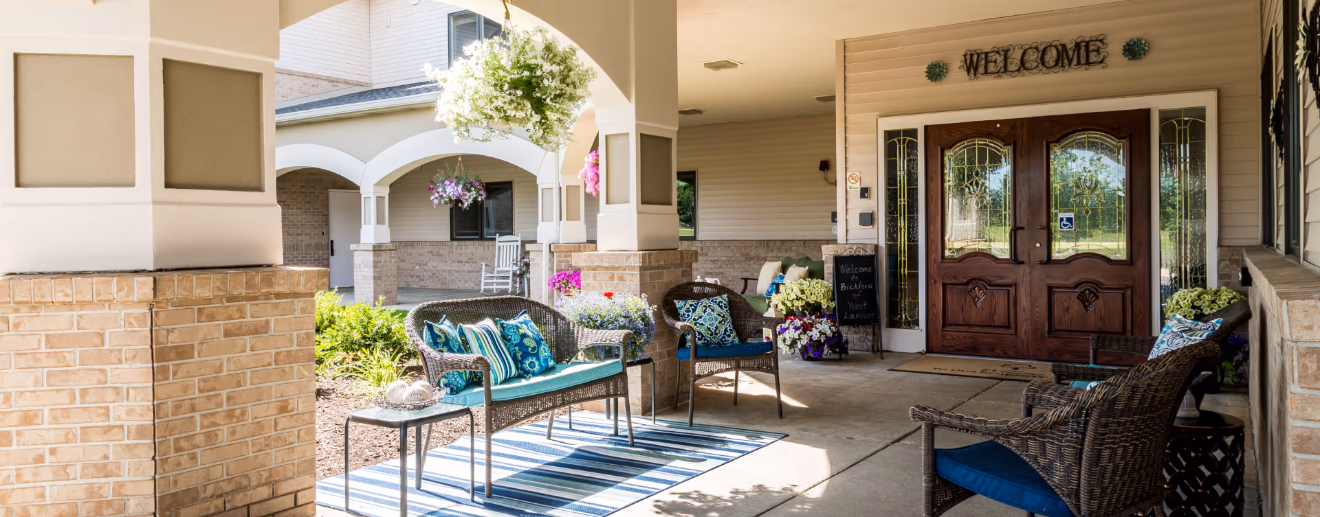 Covered outdoor seating area at the entrance of a building with wicker chairs and a bench with colorful cushions, a striped rug, hanging flower baskets, potted plants, and a wooden double door with a 'WELCOME' sign above it.