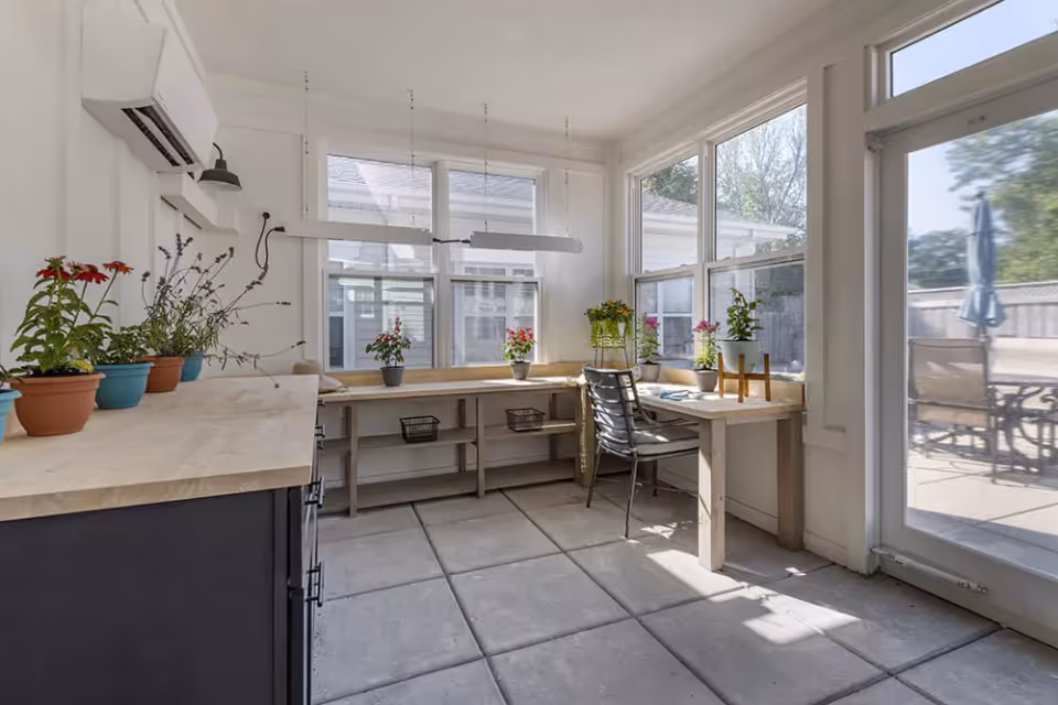 A bright indoor sunroom or workspace with large windows letting in natural light, a wooden desk with a metal chair, several potted plants on the desk and countertop, and a glass door leading to an outdoor patio area with chairs and an umbrella.