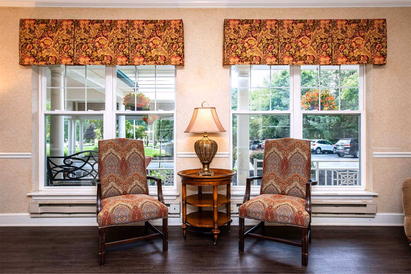 Two patterned armchairs flank a round wooden side table with a lamp in front of large windows showing a porch and parked cars outside.