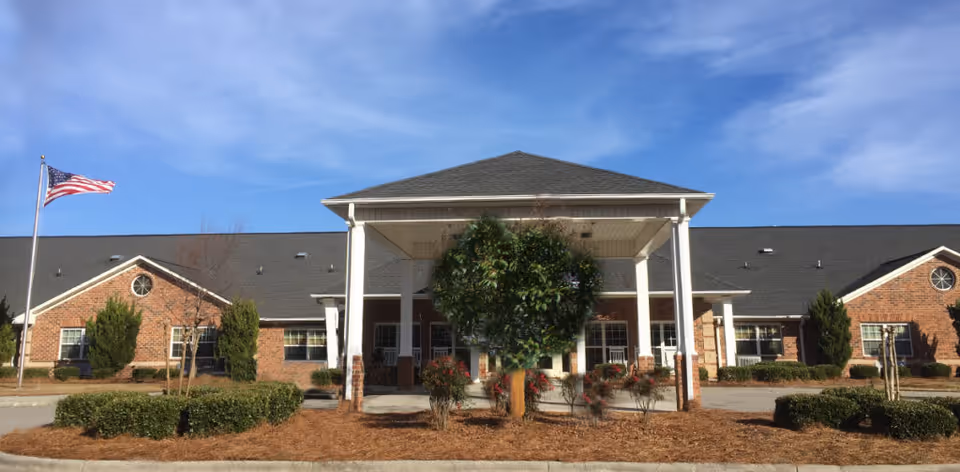 Front exterior view of a single-story brick building with a covered entrance supported by white columns. There is a small landscaped area with bushes and a tree in front, and an American flag flying on a flagpole to the left. The sky is clear and blue.