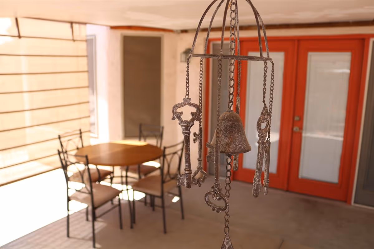 A close-up view of a decorative metal wind chime featuring keys and a bell hanging in an outdoor covered patio area. In the background, there is a round wooden table with four metal chairs and bright red double doors with glass panels.
