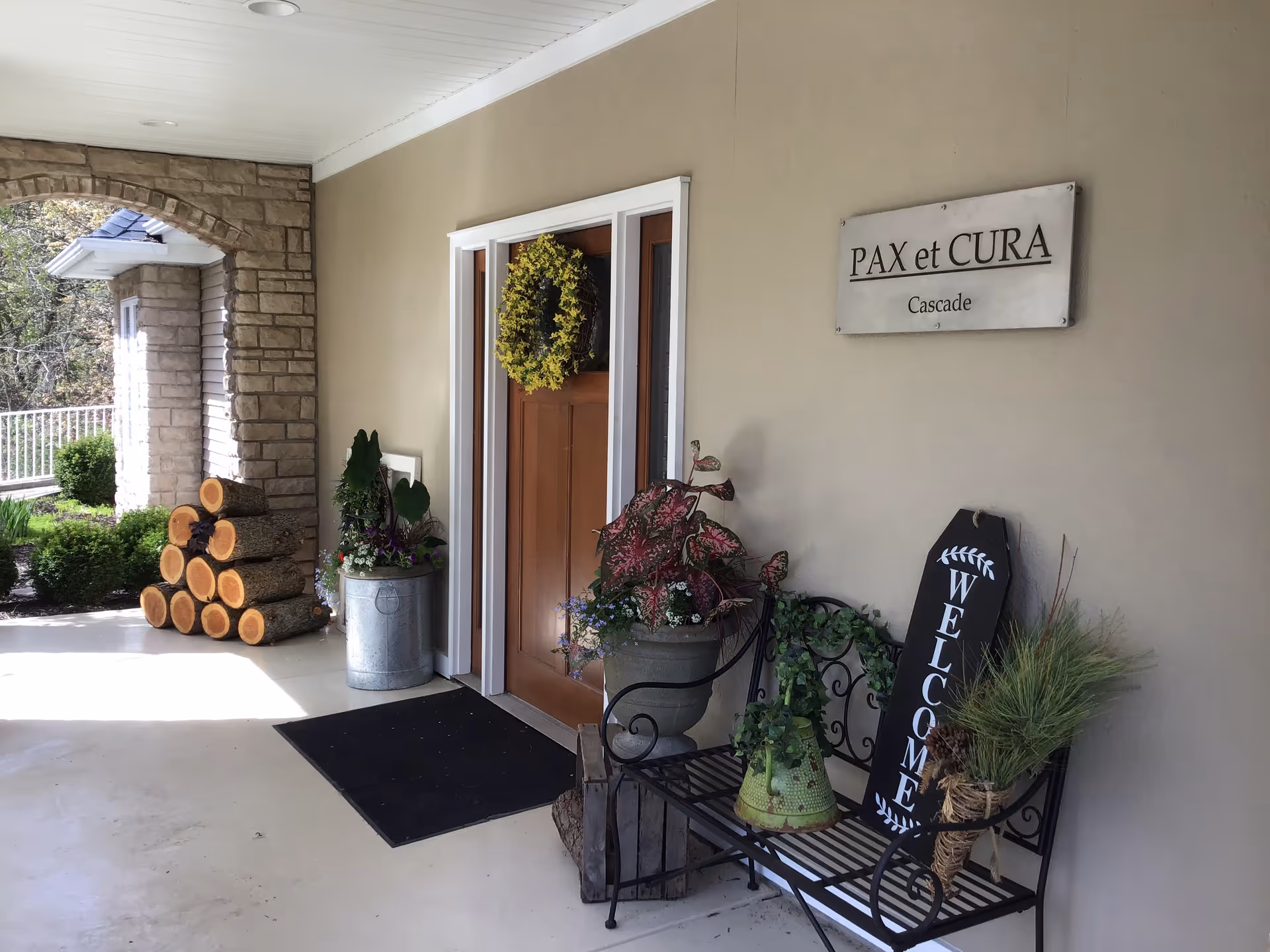 Covered entrance to Pax et Cura of Cascade showing a door with a wreath, potted plants, a bench with a 'WELCOME' sign, stacked firewood, and a wall sign reading 'PAX et CURA Cascade'.