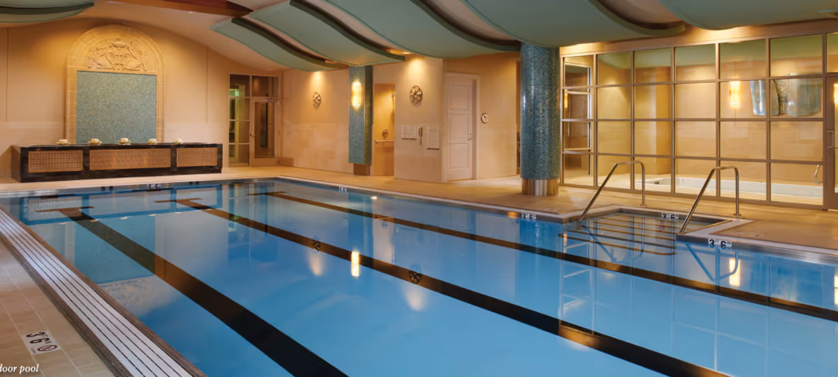 Indoor swimming pool with clear blue water, black lane markings, and steps leading into the pool. The pool area features beige tiled walls, decorative blue mosaic columns, and a water feature on one wall. The ceiling has a wave-like design with soft lighting, and there is a glass partition separating the pool from another room.