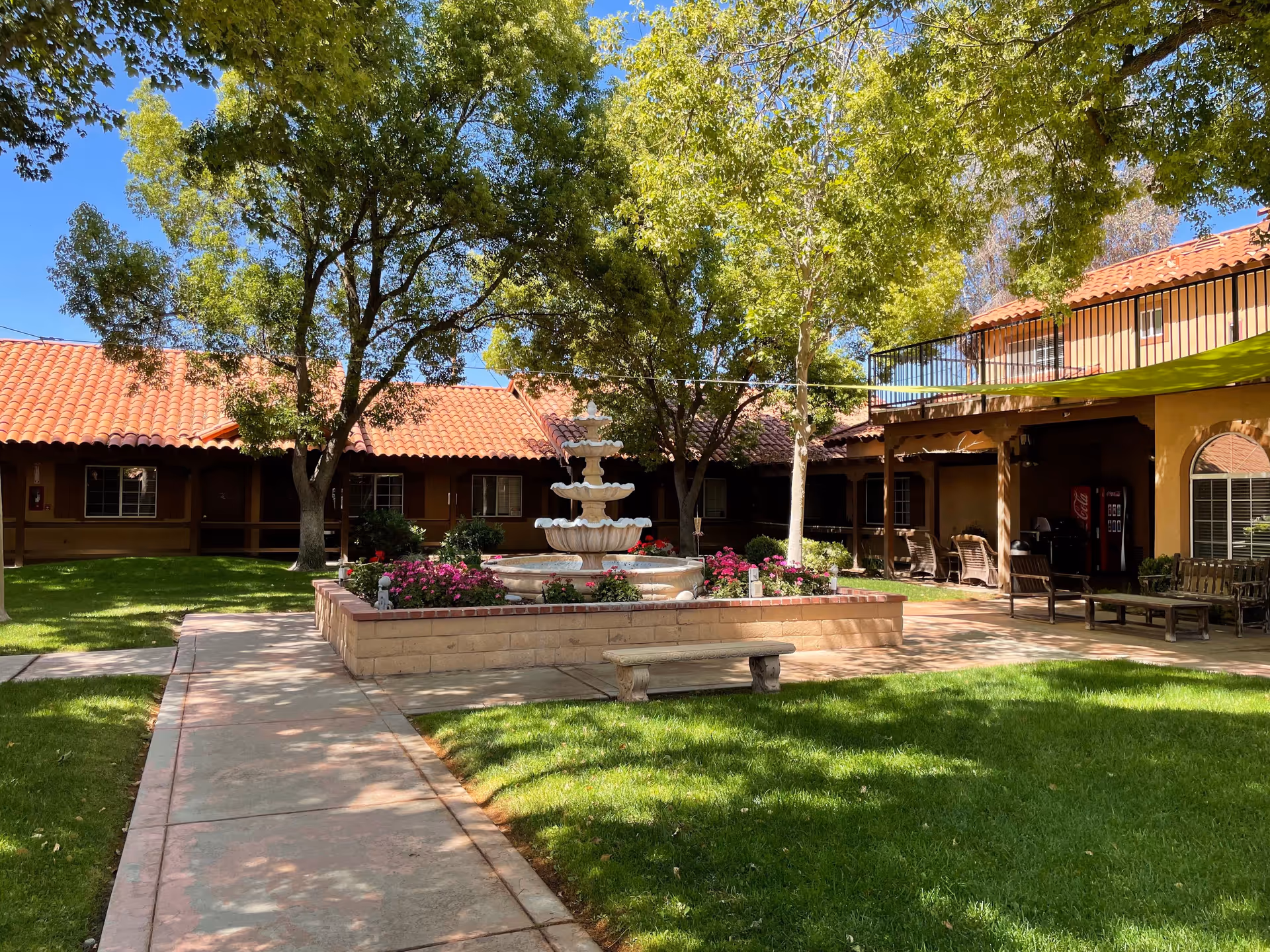 Outdoor courtyard area at Hacienda Senior Living with a central multi-tiered fountain surrounded by flower beds, green grass, trees providing shade, and a walkway leading through the space. The courtyard is bordered by a building with a red tile roof and shaded seating areas.