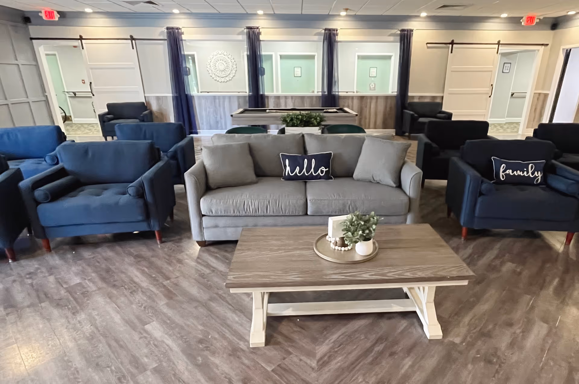 A cozy living room area with a gray sofa and two blue armchairs, each with decorative pillows that say 'hello' and 'family'. A wooden coffee table with a small plant and decorative items is in front of the sofa. The room has wood flooring, light-colored walls, and windows with dark curtains. There are doors and an exit sign visible in the background.