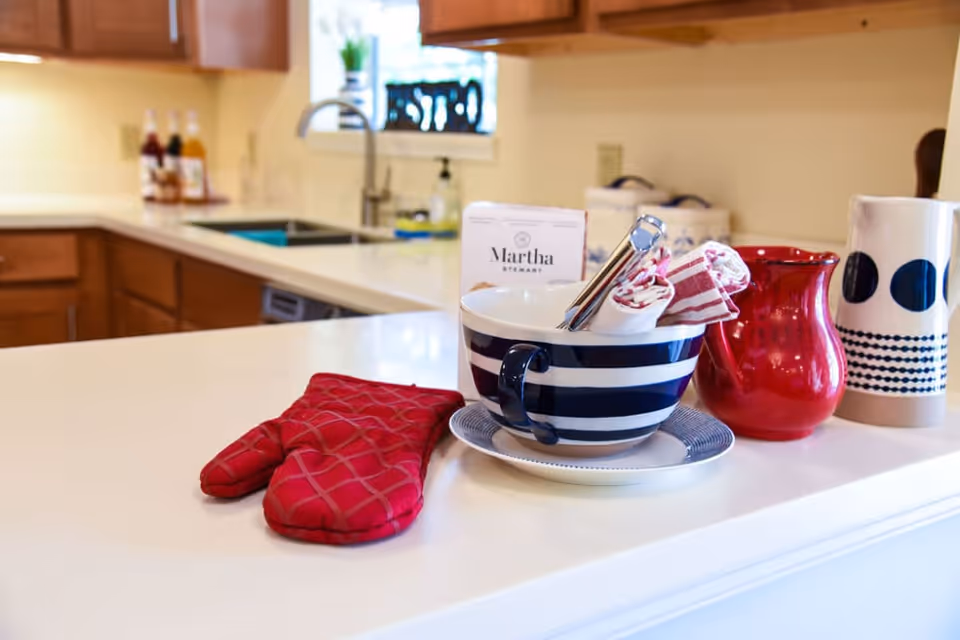 A kitchen countertop with a red oven mitt, a large striped bowl containing kitchen utensils and rolled cloth napkins, a red pitcher, and a white pitcher with blue patterns. In the background, there are wooden cabinets, a sink, and bottles on the counter near a window.