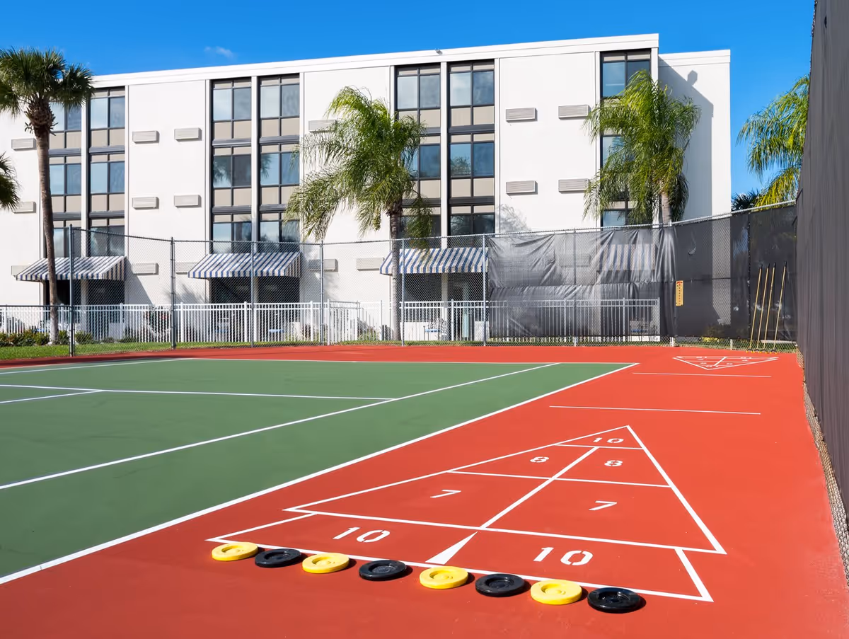 Outdoor shuffleboard court with red and green playing surface, yellow and black shuffleboard discs lined up at the start. In the background, there is a white multi-story building with large windows, palm trees, and striped awnings over the entrances. The court is enclosed by a chain-link fence with a privacy screen on one side.