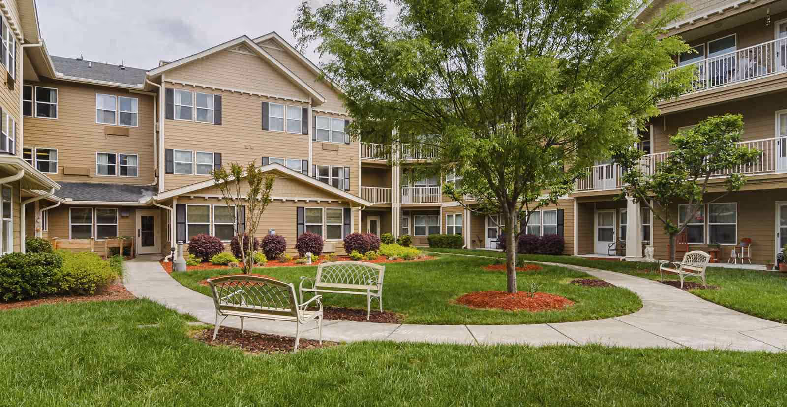 Well-maintained courtyard with benches, winding walkways, a central tree and landscaped lawns surrounded by a multi-story senior living building.
