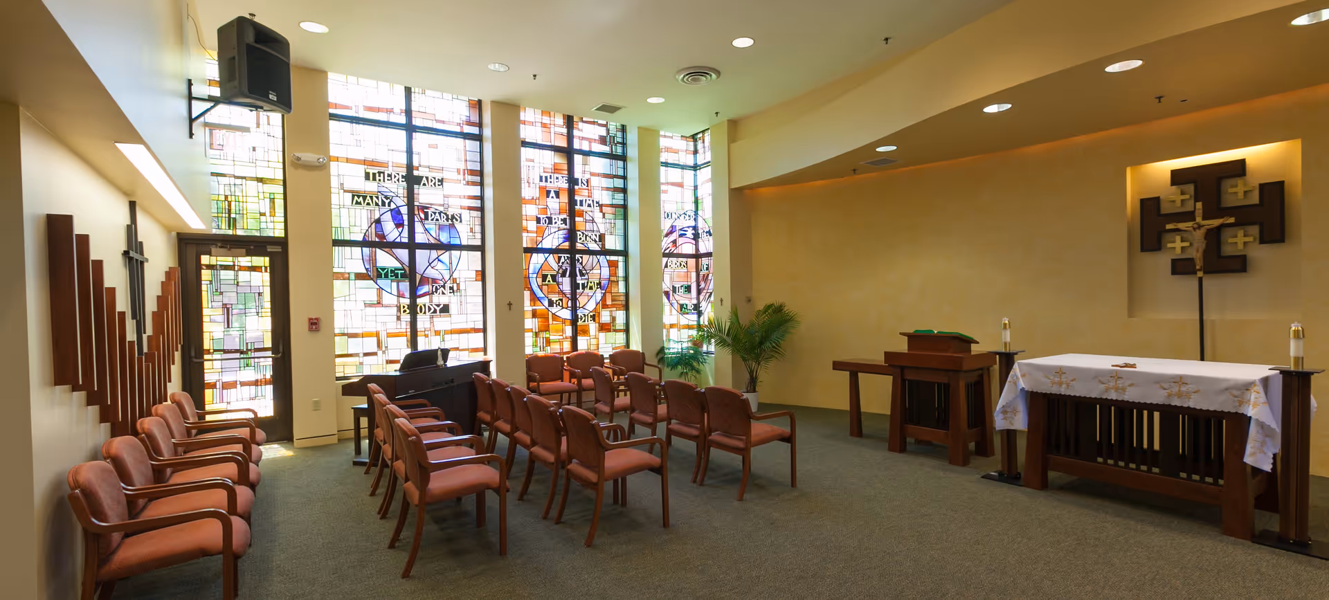 Interior view of a chapel with rows of wooden chairs with padded seats arranged facing an altar. The altar is covered with a white cloth and has candles on either side. Behind the altar is a decorative cross mounted on the wall. Large stained glass windows with colorful designs and religious text allow natural light to fill the room. There is a piano near the windows and a small wooden podium to the right of the altar.