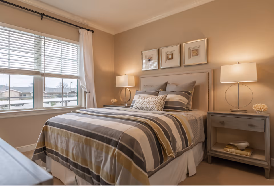 A neatly made bed with striped bedding in shades of gray, white, and beige in a bedroom. The bed has multiple pillows and a beige upholstered headboard. On either side of the bed are matching gray nightstands with modern lamps and decorative items. A large window with white blinds and curtains lets in natural light, and three framed artworks hang above the headboard on a beige wall.