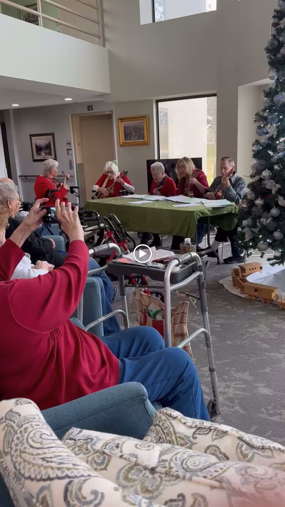 A group of elderly people seated in a common area, some playing string instruments at a table covered with a green cloth. Other residents are seated nearby, watching and taking photos. A decorated Christmas tree is visible on the right side of the room.