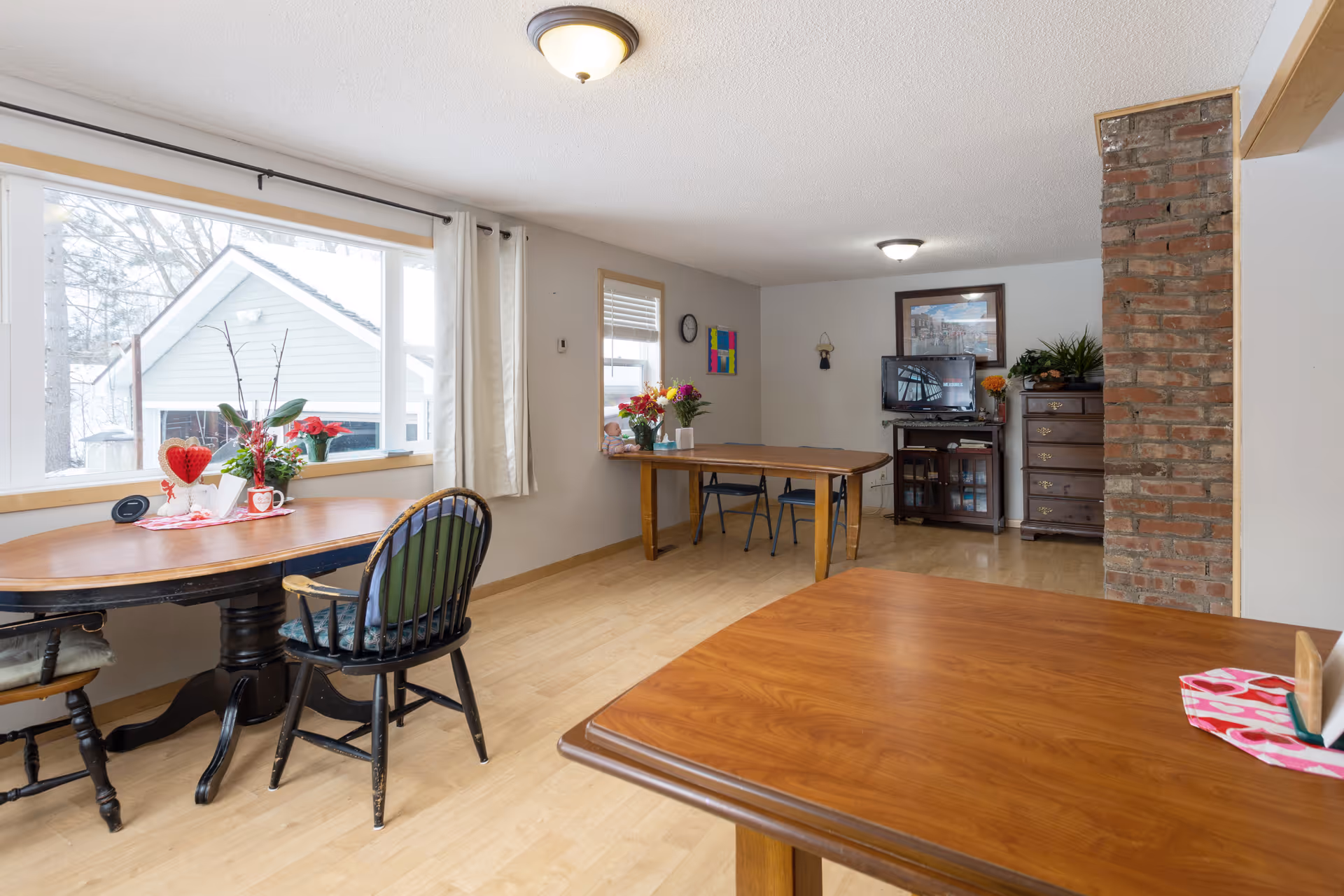 A bright room with wooden tables and chairs, decorated with flowers and a heart-shaped ornament on one table. There is a large window with white curtains letting in natural light, a brick column, a TV on a stand, and a chest of drawers against the wall.